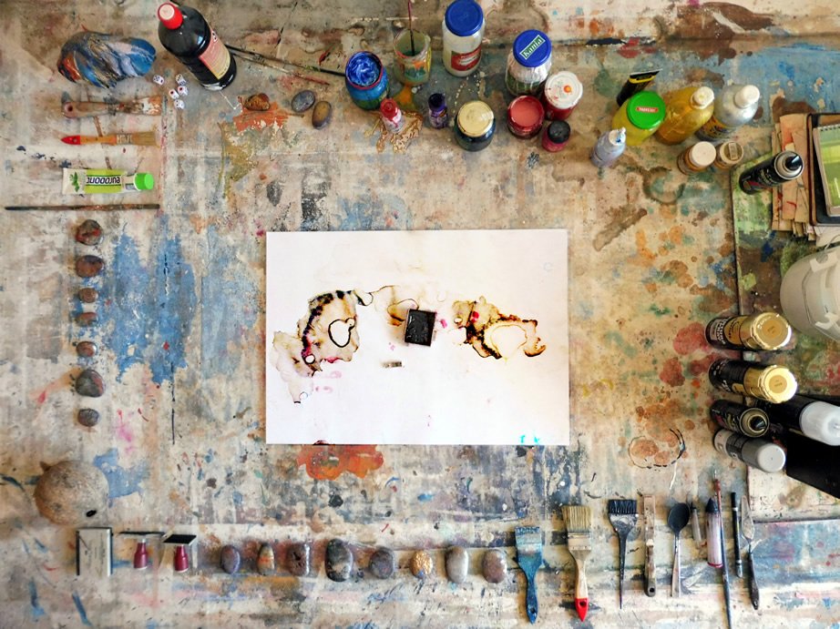 An overhead studio photograph showing brushes, pigments, materials and a work on paper